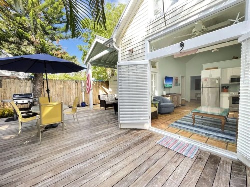 A sunny outdoor patio with a dining set, grill, and view of a cozy living area through open doors.