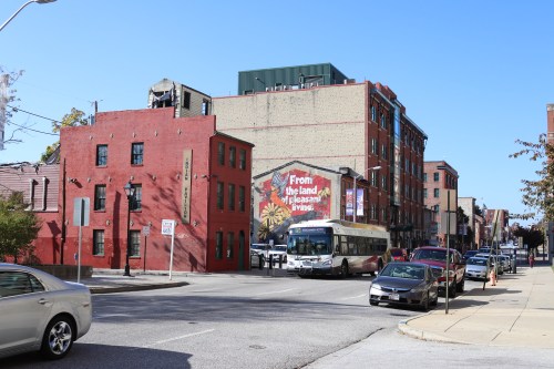 A city street scene featuring red buildings, a bus, and a mural on a brick wall under a clear blue sky.