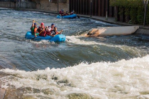 A group of people in a blue raft navigating a river with rapids, surrounded by greenery and a stone wall.