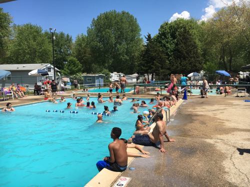 A crowded outdoor pool scene with people swimming, lounging, and enjoying a sunny day under blue skies.