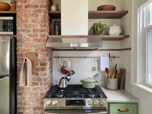 Cozy kitchen corner with a gas stove, brick wall, open shelves, and various cooking utensils and plants.