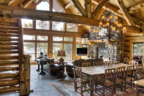 Cozy log cabin interior with a large dining table, stone fireplace, and large windows showcasing a scenic view.