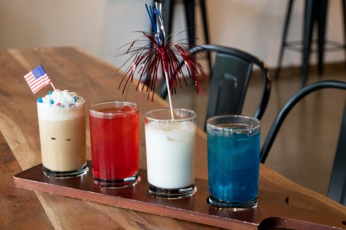 Four colorful drinks on a wooden tray: a coffee drink, red soda, white creamy drink, and blue beverage, decorated for a celebration.
