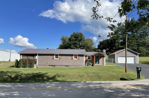 A single-story house with a gray exterior, green lawn, and a detached garage under a blue sky with clouds.