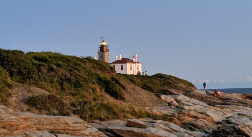 A lighthouse on a rocky shore, surrounded by greenery, with people walking along the coast under a clear blue sky.