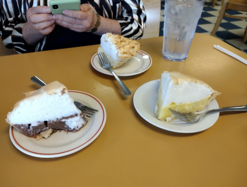 Three slices of cake on a table: chocolate, lemon meringue, and coconut meringue, with forks and a glass of water.