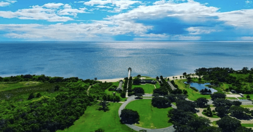 Aerial view of a coastal landscape featuring a lighthouse, green parks, and a calm blue sea under a partly cloudy sky.