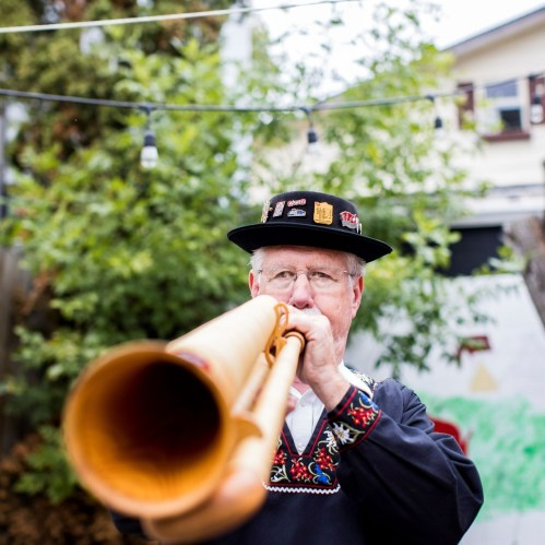 An older man in traditional attire plays a wooden horn in a garden setting, surrounded by greenery.