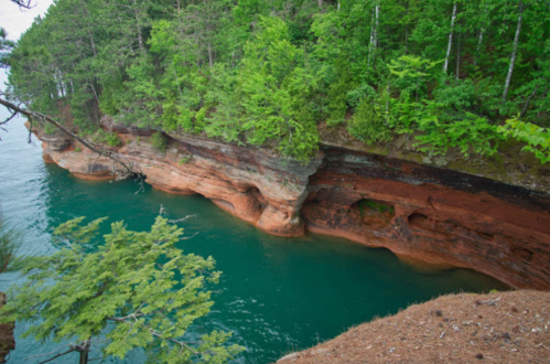 A scenic view of a rocky shoreline with green trees and clear blue water, showcasing layered rock formations.