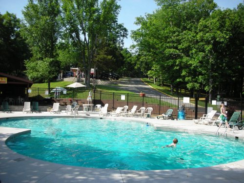 A sunny outdoor pool area surrounded by lounge chairs and trees, with people swimming and a playground in the background.