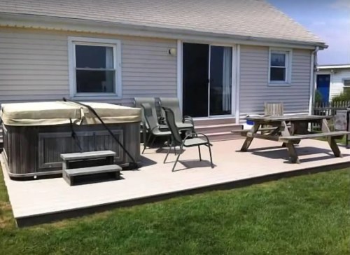 A backyard patio with a hot tub, chairs, and a picnic table, next to a house with sliding glass doors.