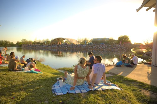 A couple sits on a blanket by a lake, enjoying a sunset while a crowd gathers for an outdoor event in the background.