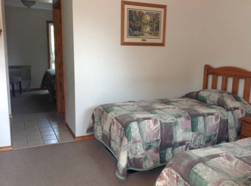 A simple bedroom with two twin beds, a wooden headboard, and a doorway leading to another room.