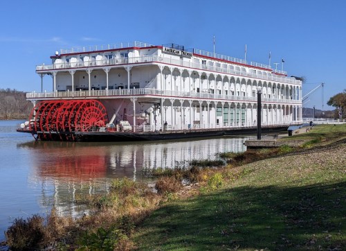 A large white riverboat with red paddle wheels docked by a river, surrounded by greenery and a clear blue sky.