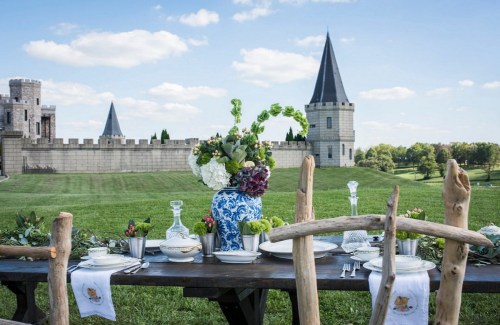 A beautifully set outdoor dining table with flowers, overlooking a castle-like structure and green landscape.