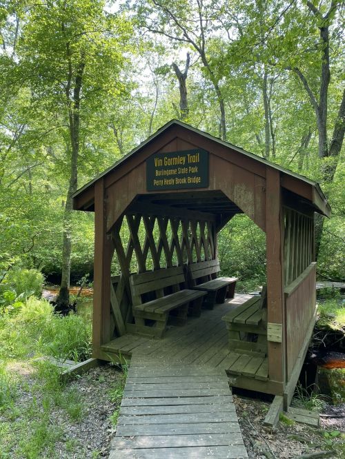 A wooden shelter on the Vin Gormley Trail in a lush green park, with benches and a sign for Perry Helvy Brook Bridge.