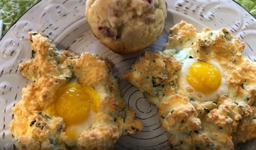 Two baked eggs in fluffy green herb clouds on a plate, alongside a muffin with visible fruit pieces.