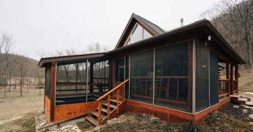 A rustic wooden cabin with screened porches, surrounded by trees and a grassy area, set against a cloudy sky.