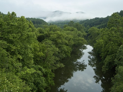 Lush green trees line a calm river, reflecting the overcast sky and distant mountains shrouded in mist.