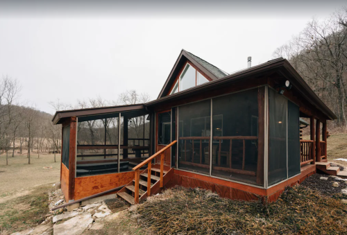A rustic cabin with large screened porches, surrounded by trees and grassy land, set against a hilly backdrop.