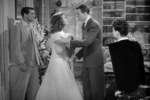A tense moment in a vintage living room with a man and woman in formal attire, while two others observe.