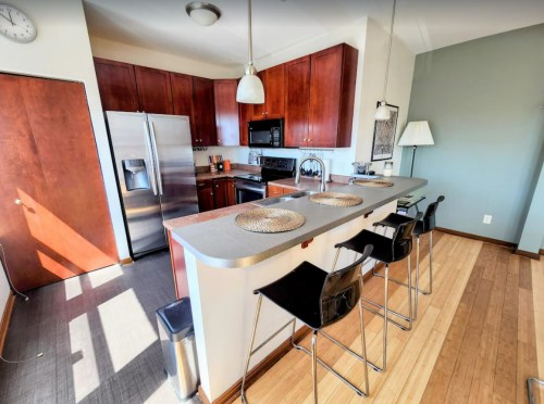 Modern kitchen with wooden cabinets, stainless steel appliances, and a breakfast bar with black stools. Bright and spacious.