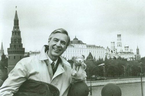 A smiling man in a coat holds a stuffed animal, with a cityscape featuring towers and buildings in the background.