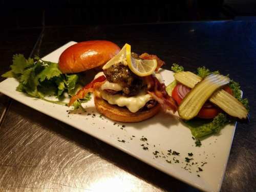 A gourmet burger topped with bacon, cheese, and lemon, served with lettuce, pickles, and tomato on a white plate.