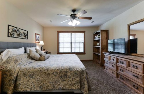 Cozy bedroom with a large bed, ceiling fan, TV, and wooden furniture, featuring natural light from a window.