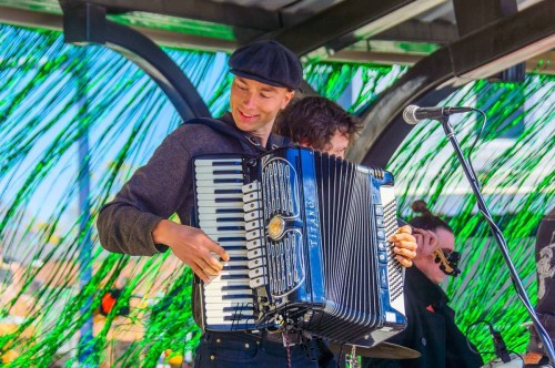 A musician plays an accordion on stage, smiling, with a colorful backdrop of green and orange.