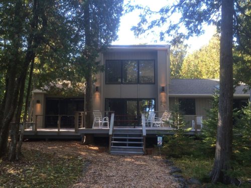A modern house surrounded by trees, featuring a large deck and multiple windows, set in a natural landscape.
