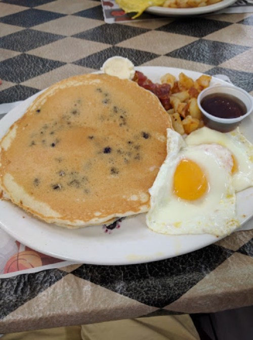 A plate of breakfast featuring a large blueberry pancake, fried eggs, hash browns, and a small cup of syrup.