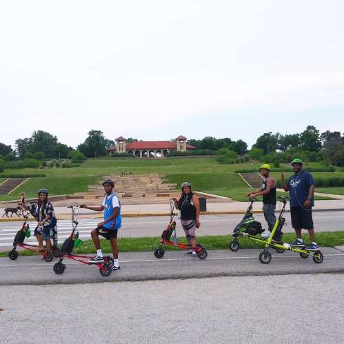 A group of five people riding scooters on a path, with a park and building in the background.