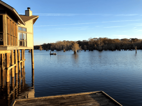 A serene lake view with a wooden house on stilts, surrounded by trees and a small boat in the distance under a clear sky.