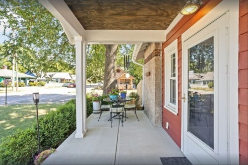 A cozy porch with a table and chairs, overlooking a green yard and street with trees and houses in the background.