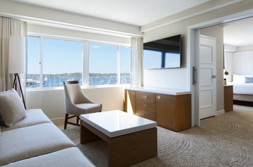 Bright hotel room with a view of the marina, featuring a sofa, coffee table, TV, and a door leading to a bedroom.