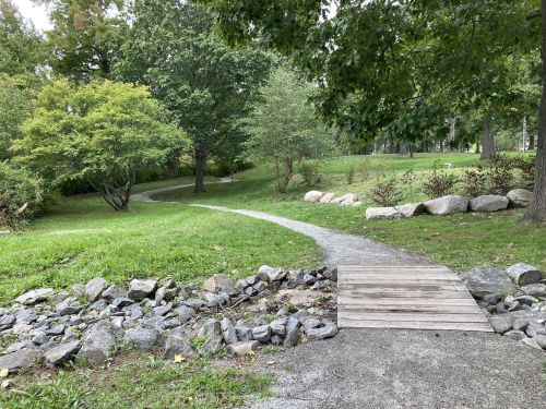 A winding path through a green park, with a wooden bridge over rocks and trees lining the area.
