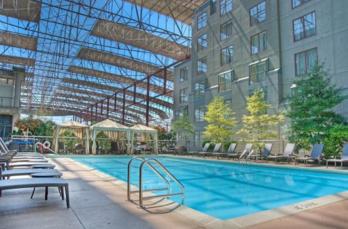 A bright indoor pool area with lounge chairs, surrounded by greenery and a modern building structure overhead.