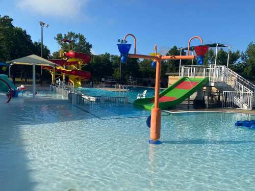 A sunny pool area featuring water slides, splash features, and lounge chairs surrounded by trees.