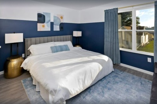 A modern bedroom with a large bed, blue accent wall, two lamps, and a window showing an outdoor view.