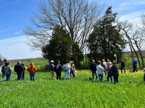 A group of people stands in a green field under a blue sky, with trees in the background.