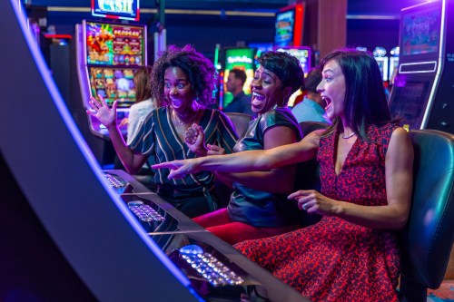 Three women excitedly play slot machines in a vibrant casino, celebrating a win with joyful expressions.