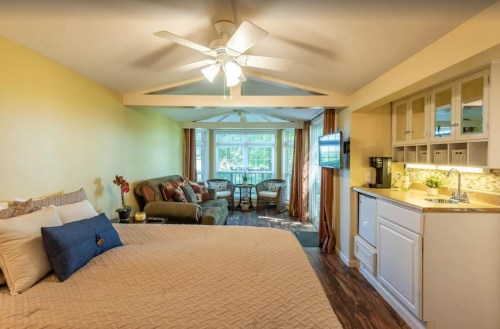 Cozy room with a bed, seating area, and kitchenette, featuring large windows and warm yellow walls.