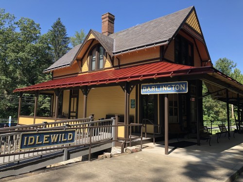 Historic train station with yellow and red exterior, featuring signs for "Idlewild" and "Darlington" surrounded by trees.