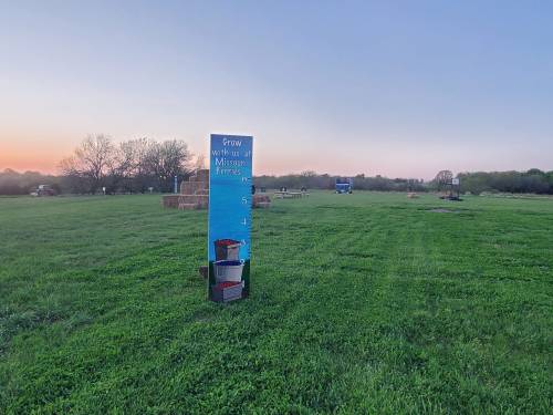 A sign in a grassy field at sunset, with hay bales and a blue tractor in the background.