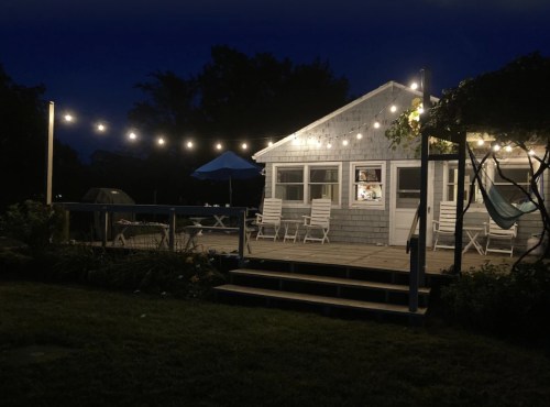 A cozy house at night, illuminated by string lights, with a deck and outdoor seating.