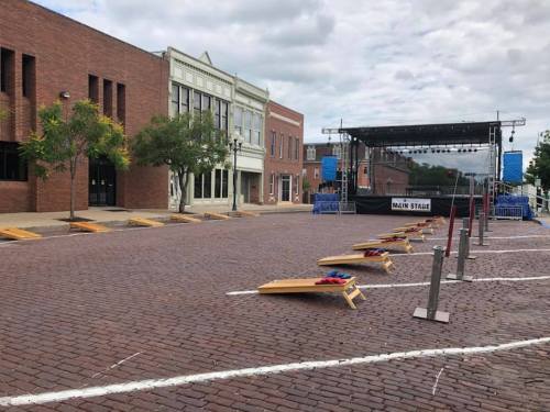 Empty street with cobblestones, featuring a stage and cornhole boards set up for an event. Overcast sky above.