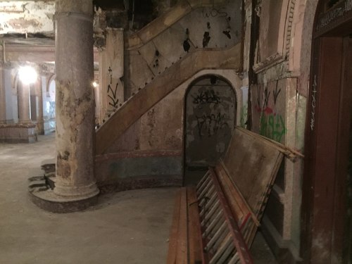 Abandoned interior with a staircase, graffiti on walls, and old wooden benches in a dimly lit space.