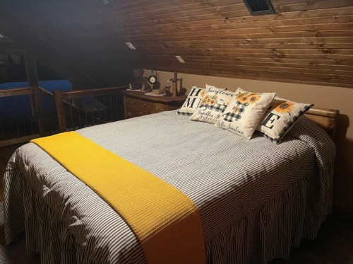 Cozy bedroom with a wooden ceiling, a large bed dressed in striped bedding, and decorative pillows. A yellow throw blanket adds color.