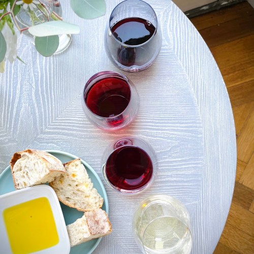 Three glasses of red wine and one glass of white wine on a textured table, accompanied by bread and olive oil.